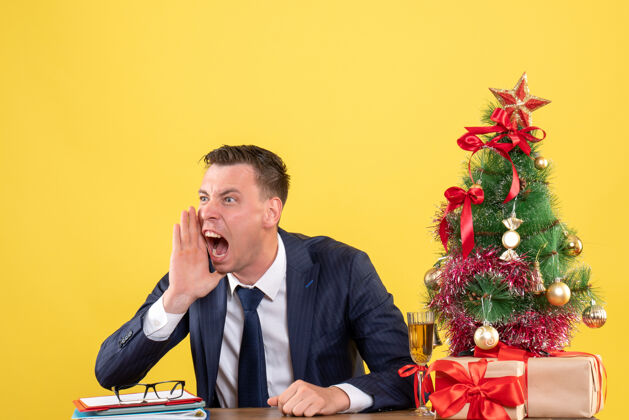 男性西装革履的男人坐在圣诞树旁的桌子旁大声喊叫 黄色的礼物圣诞节西装新郎