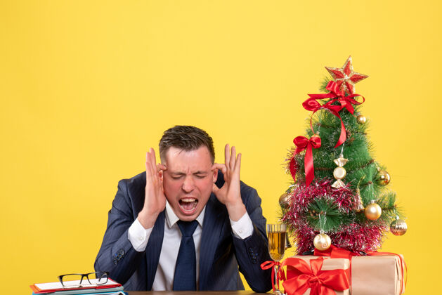 男人正面图：一个男人举着耳朵坐在圣诞树旁的桌子上 黄色的礼物坐着抱着办公室
