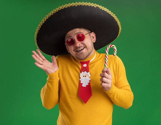 手杖穿着黄色高领毛衣 戴着眼镜 打着滑稽的红领带 戴着黑色帽子 手里拿着糖果棒 脸上带着幸福的笑容站在绿色的墙上帽子高领毛衣领带