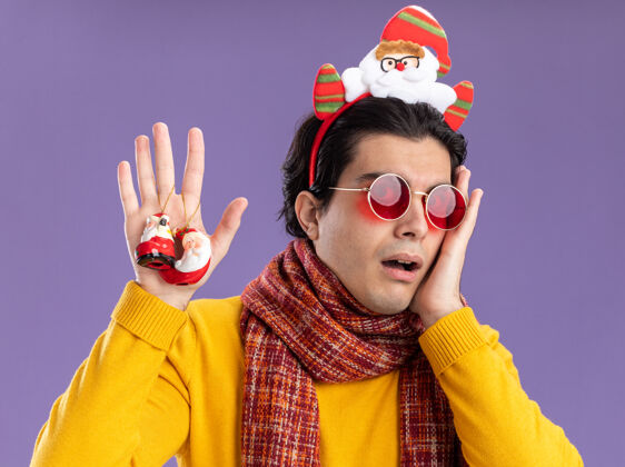 年轻脖子上围着暖围巾 穿着黄色高领毛衣 头上戴着滑稽的眼镜 手里拿着圣诞玩具 忧心忡忡地站在紫色的墙上边缘高领毛衣玩具