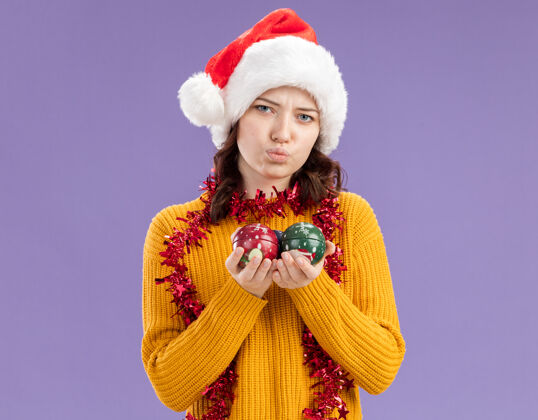 球失望的年轻斯拉夫女孩戴着圣诞帽 脖子上戴着花环 手里拿着玻璃球饰物 背景是紫色 有复制空间装饰品女孩周围