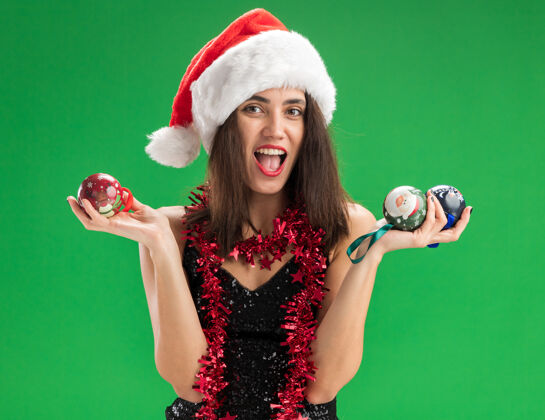 球快乐的年轻漂亮的女孩戴着圣诞帽 脖子上戴着花环 手里拿着圣诞树上的球 隔离在绿色的墙上帽子快乐女人