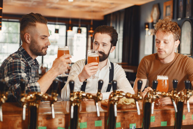 酒吧和最好的朋友见面三个穿着休闲服的快乐的年轻人一起坐在酒吧里聊天喝啤酒朋友胡须会议