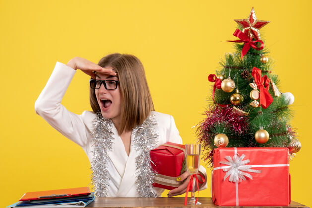 现在正面图：女医生坐在桌子前 手里拿着黄色背景的礼物 还有圣诞树和礼品盒桌子人抱