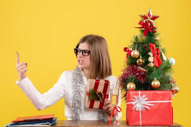 女性正面图：女医生坐在桌子前 手里拿着黄色背景的礼物 还有圣诞树和礼品盒人快乐新的