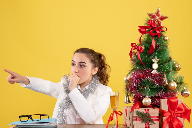医生前视图：女医生坐在桌子后面 惊恐地指着黄色背景上的圣诞树和礼品盒坐着前面女