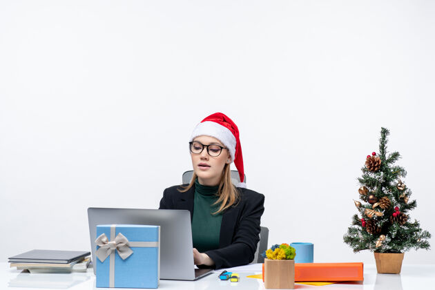 工作戴着圣诞老人帽的女商人坐在桌子旁 桌上放着圣诞树和礼物笔记本电脑办公室圣诞树