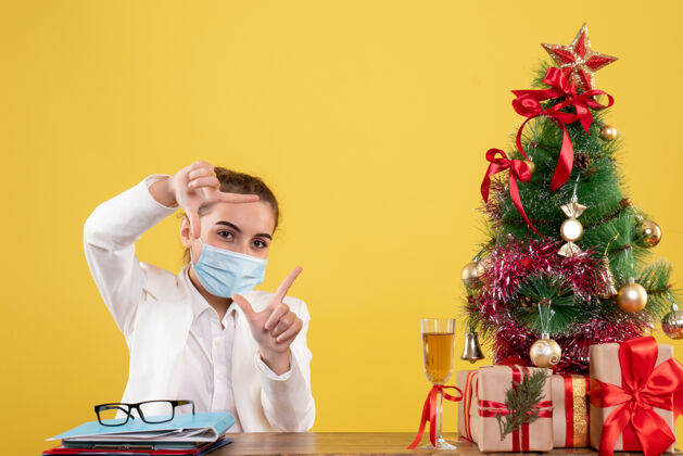 脸正面图女医生戴着黄色背景的无菌口罩坐在圣诞树和礼盒里快乐坐着女性
