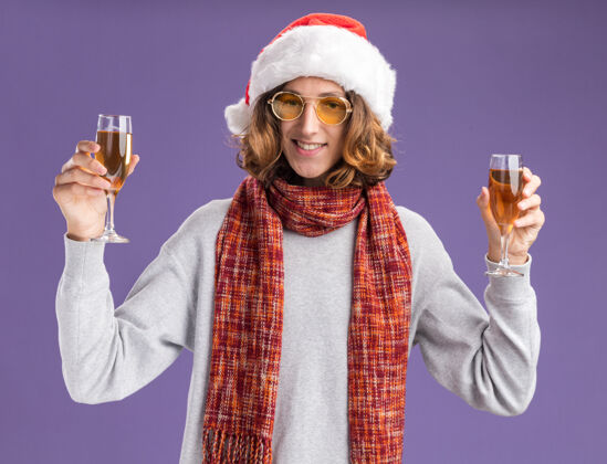 微笑快乐的年轻人戴着圣诞老人的帽子 戴着黄色的眼镜 脖子上围着暖和的围巾 手里拿着香槟酒杯 站在紫色的背景下兴高采烈地微笑着看着相机香槟脖子看着