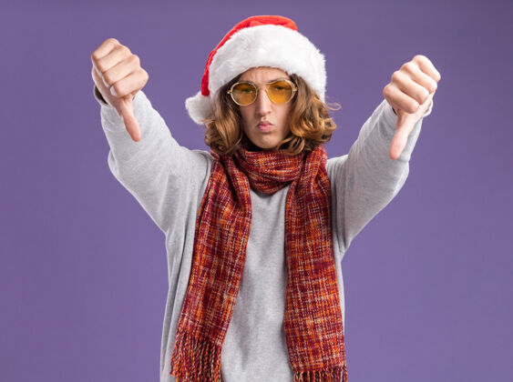拇指一个戴着圣诞圣诞老人帽 戴着黄色眼镜 脖子上围着暖和的围巾的年轻人 严肃地皱着眉头 站在紫色的背景上竖起大拇指 看着摄像机帽子围巾严重