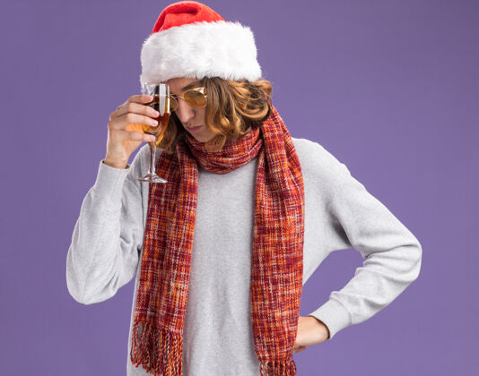 抱着年轻人戴着圣诞老人帽 戴着黄色眼镜 脖子上围着一条暖和的围巾 手里拿着一杯香槟 站在紫色的背景下 看上去又累又无聊 很沮丧香槟圣诞节站着