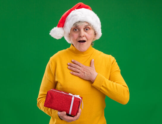 胸前兴奋的老妇人戴着圣诞帽 把手放在胸前 拿着一个紫色背景的圣诞礼盒 上面留有复制空间举行老人新