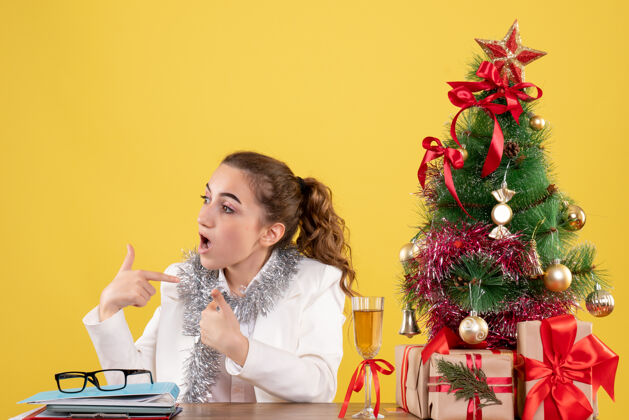 坐前视图：女医生坐在桌子后面 拿着圣诞礼物和黄色背景上的树漂亮肖像快乐