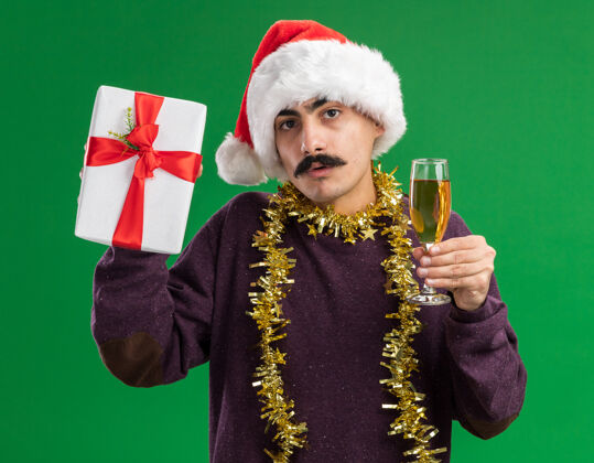 圣诞一个留着胡子的年轻人戴着圣诞老人帽子 脖子上戴着金属丝 手里拿着一杯香槟和圣诞礼物 站在绿色的背景下困惑地看着摄像机快乐圣诞老人帽子