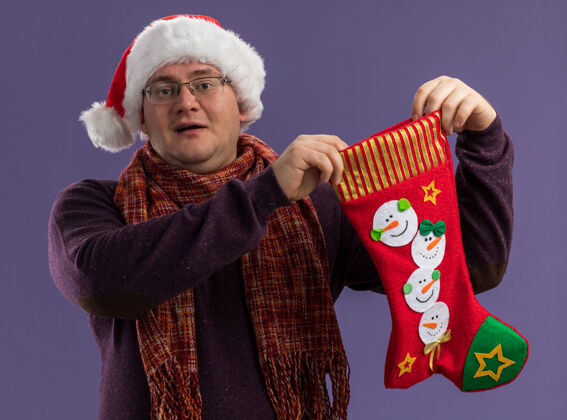 抱着给人印象深刻的成年男子戴着眼镜 戴着圣诞帽 脖子上围着围巾 手里拿着圣诞长袜 看着隔离在紫色背景上的相机圣诞老人围着脖子