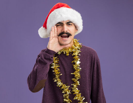 圣诞年轻的胡子男人戴着圣诞老人帽子 脖子上戴着金属丝 手靠近嘴 站在紫色的背景上大声喊叫周围圣诞老人年轻