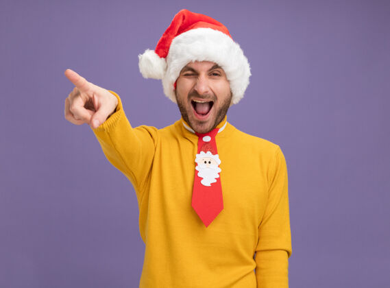 圣诞快乐自信的年轻白种人戴着圣诞帽打着领带看着摄像机眨着眼睛指着一边张大嘴巴隔离在紫色背景上 留着复印空间帽子自信圣诞节