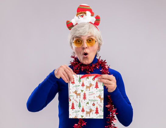 圣诞老人戴着太阳眼镜 脖子上戴着圣诞老人头带和花环 手里拿着纸质礼品袋的兴奋的老妇人眼镜花环年