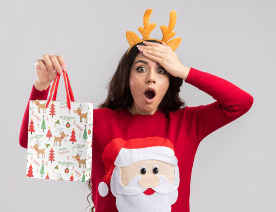 漂亮遗憾的是年轻漂亮的女孩戴着驯鹿鹿角头带和圣诞老人毛衣拿着圣诞礼物包看着手放在额头上头带快乐手