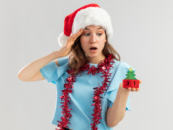 年身穿蓝色上衣 戴着圣诞帽 脖子上戴着金属丝的年轻女子 手里拿着玩具立方体 与新年快乐约会 看着立方体 惊讶不已圣诞老人玩具惊奇