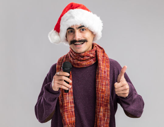 脖子年轻的留着胡子的男子戴着圣诞老人帽 脖子上围着暖和的围巾 手里拿着麦克风 面带微笑 高兴地竖起大拇指展示站着戴着