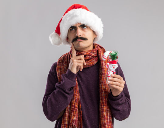 站着年轻的胡子男人戴着圣诞老人帽 脖子上围着暖和的围巾 手里拿着圣诞糖果手杖 疑惑地抬起头来年轻人抱着戴着