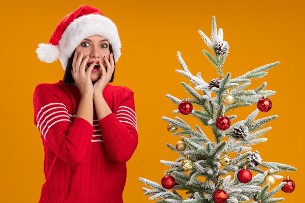 圣诞老人令人印象深刻的年轻女孩戴着圣诞帽站在装饰圣诞树附近 手放在橙色墙上的脸隔离印象深刻靠近年轻
