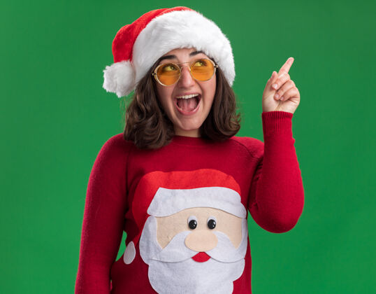 穿着穿着圣诞毛衣戴着圣诞帽戴着眼镜的小女孩抬起头 高兴地用食指指着绿色的墙壁帽子手指食指