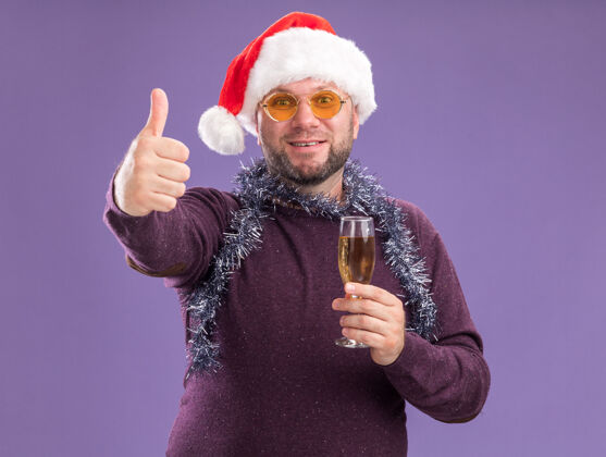 香槟微笑的中年男子戴着圣诞老人帽 脖子上戴着金箔花环 戴着眼镜 手里拿着一杯香槟 在紫色的墙上孤立地竖起大拇指圣诞老人脖子中年