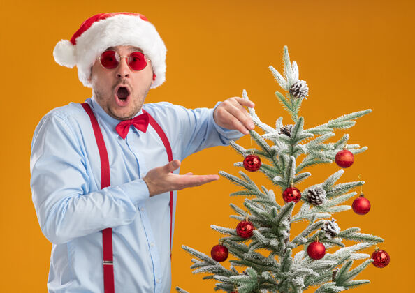 圣诞老人那个戴着圣诞帽 戴着红眼镜 系着吊带领结的年轻人站在圣诞树旁 高兴而惊奇地把玩具挂在橘色墙上的树上领带圣诞节吊带