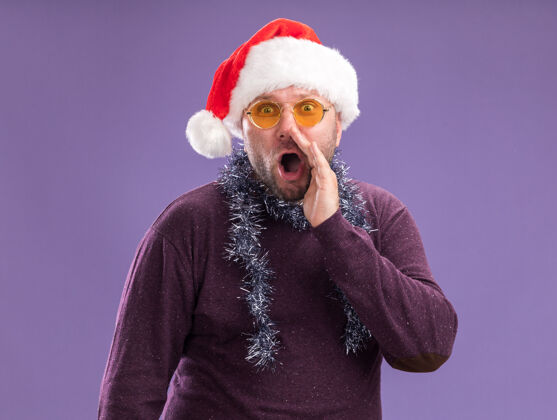 金属丝令人印象深刻的中年男子戴着圣诞帽 脖子上戴着金箔花环 戴着眼镜 在紫色的墙上低语着周围穿着中年