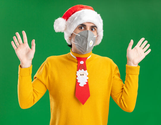保护穿着黄色高领毛衣 戴着圣诞帽 打着滑稽领带 戴着护面面具的年轻人担心地举起双臂站在绿色的墙上提高圣诞节帽子