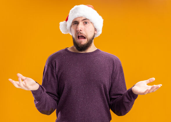 男人身穿紫色毛衣 头戴圣诞帽的年轻人困惑地 不高兴地张开双臂站在橙色的墙上蔓延圣诞节圣诞老人