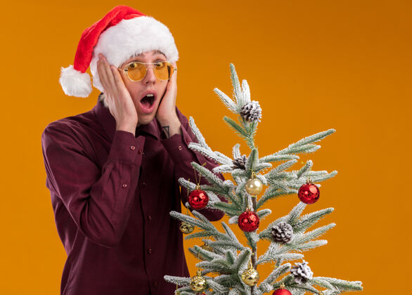 圣诞老人一个戴着圣诞帽和眼镜的年轻金发男人站在装饰好的圣诞树旁 手放在橘色的墙上 脸上与世隔绝保持男人年轻