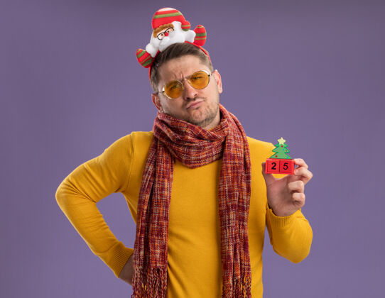 满意的年轻人穿着黄色高领毛衣 戴着暖和的围巾和眼镜 戴着滑稽的轮辋 头上戴着圣诞老人 在紫色的墙上展示着25号玩具立方体男人数字年轻