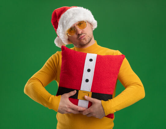 高领毛衣快乐的年轻人穿着黄色高领毛衣 戴着圣诞帽 戴着眼镜 抱着红色滑稽的枕头 脸上带着微笑站在绿色的墙上微笑年轻人圣诞老人