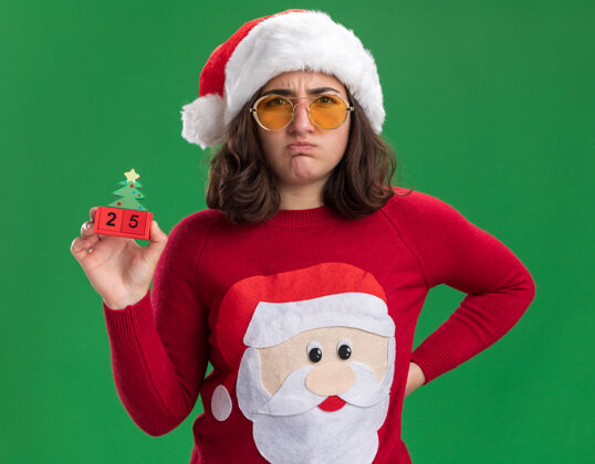噘嘴穿着圣诞毛衣 戴着圣诞帽 戴着眼镜 拿着25号玩具立方体的小女孩站在绿色的墙上 不高兴地噘着嘴唇穿着二十个不高兴