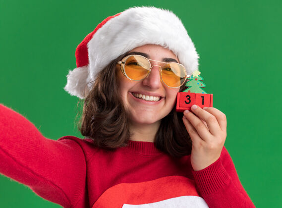 圣诞节穿着圣诞毛衣 戴着圣诞帽 戴着圣诞眼镜 手里拿着玩具方块 脸上挂着大大的笑容 站在绿色的墙上年轻方块圣诞老人