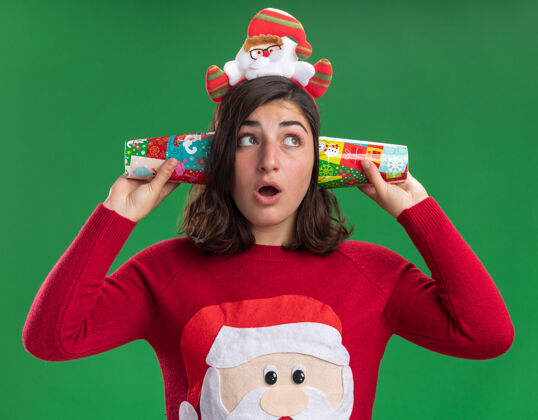 旁边穿着圣诞毛衣 戴着圣诞帽 耳边拿着五颜六色的纸杯的年轻女孩站在绿色的墙上 惊讶地看着一边圣诞老人靠近毛衣