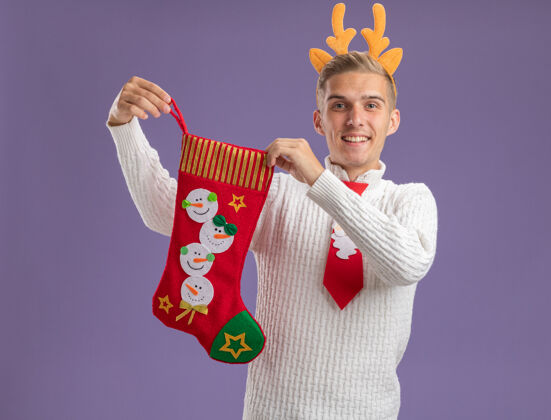 圣诞老人快乐的年轻帅哥戴着驯鹿鹿角头带 打着圣诞老人的领带 手里拿着圣诞长袜 孤零零地站在紫色的墙上年轻领带长袜