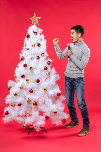 新的新年气氛积极的家伙穿着牛仔裤站在装饰圣诞树附近呼吁有人在红色雪人站着电话