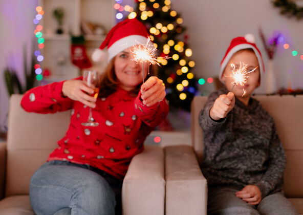 圣诞树快乐的母亲和小儿子戴着圣诞帽 戴着闪闪发光的圣诞灯 坐在沙发上 在装饰好的房间里 背景是圣诞树 尽情庆祝圣诞节小儿子快乐