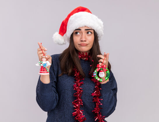 年轻失望的年轻白人女孩戴着圣诞帽 脖子上戴着花环 手里拿着圣诞树玩具 看着旁边新年新圣诞