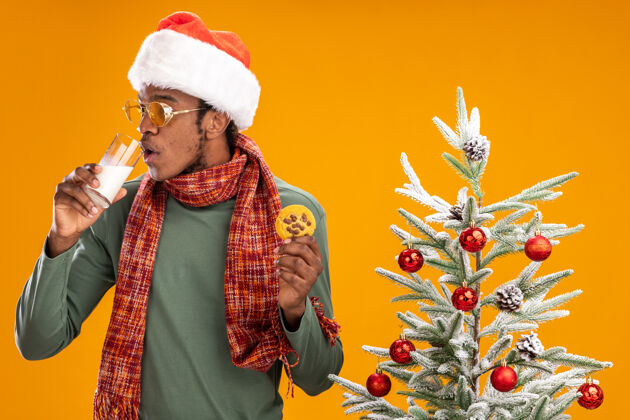 站着戴着圣诞帽 围着围巾 拿着饼干喝牛奶的非洲裔美国人站在橙色背景下的圣诞树旁喝酒牛奶圣诞树
