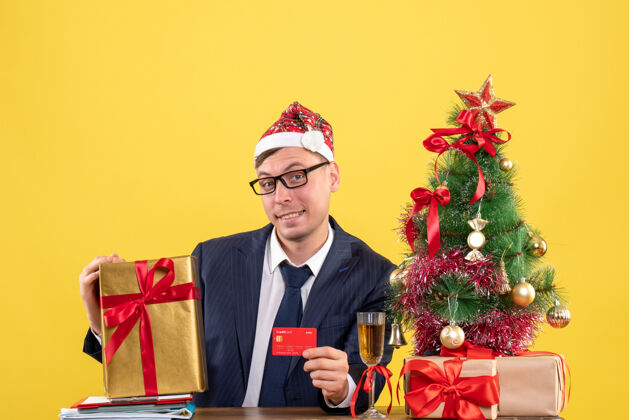 商人商务人士戴着圣诞帽坐在圣诞树旁的桌子前 黄色的礼物帽子圣诞树商务