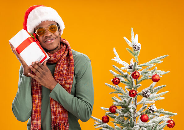 美国人戴着圣诞帽 脖子上围着围巾的非洲裔美国人拿着礼物 面带微笑地看着相机 站在橙色背景下的圣诞树旁圣诞树脖子男人