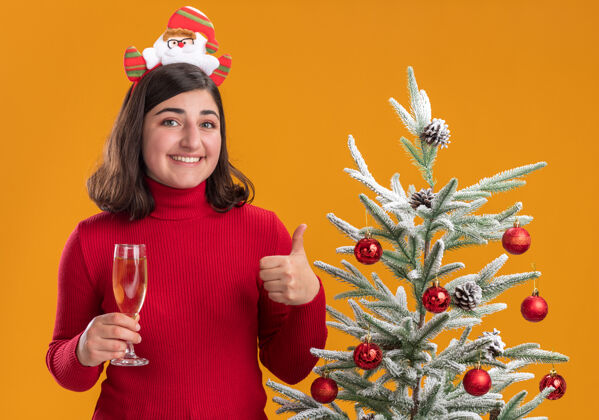 圣诞树穿着圣诞毛衣 戴着滑稽头带 手持香槟的快乐小女孩站在橙色背景下的圣诞树旁女孩玻璃杯看