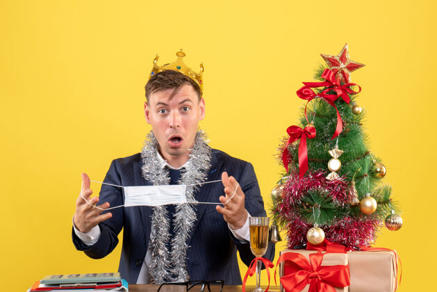 抱着前视图的商人拿着他的面具坐在圣诞树附近的桌子上 黄色的礼物面具黄色生意