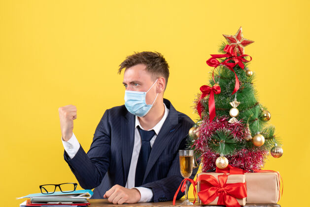 圣诞节商务人士带着医用面罩坐在圣诞树旁的桌子前 呈现黄色人树商务