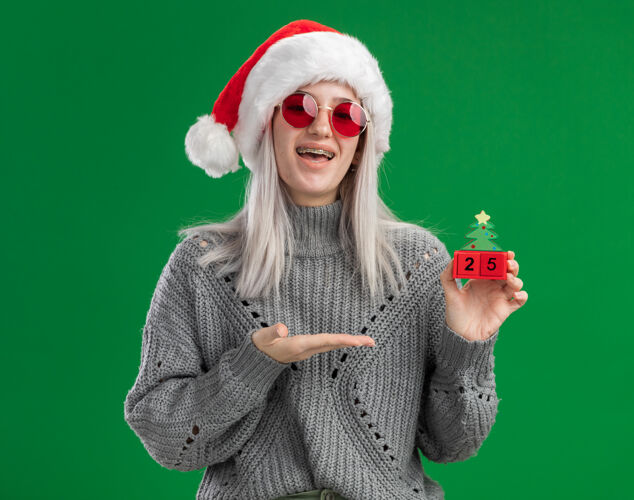金发年轻的金发女郎 穿着冬季毛衣 戴着圣诞老人帽 戴着红眼镜 抱着玩具立方体 带着圣诞节的日期 在绿色的背景下 手臂欢快而积极地站着帽子眼镜介绍